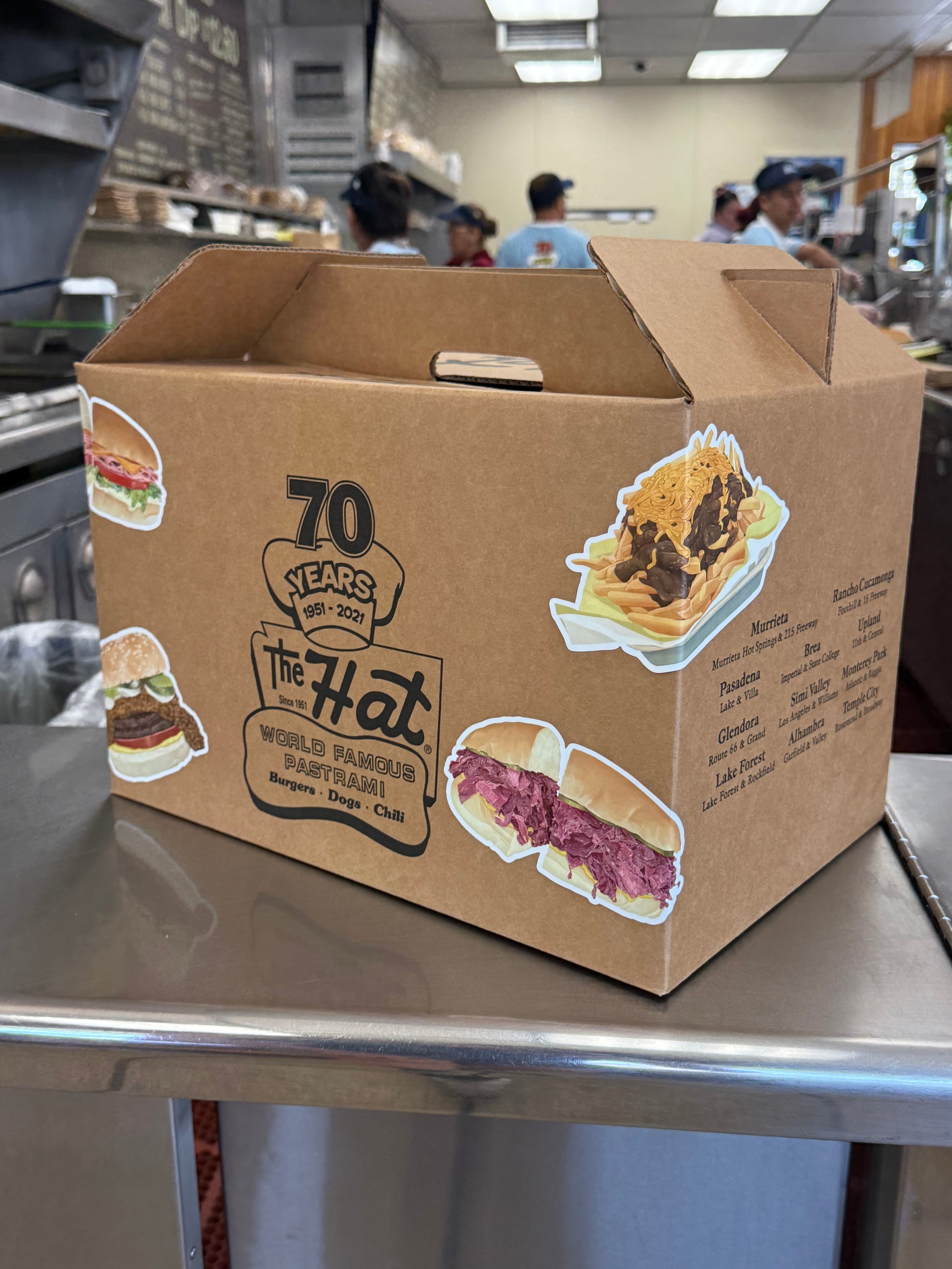 Cardboard box with food illustrations and text, held by a person in a kitchen setting. The Hat Restaurant take out box with stickers on it.